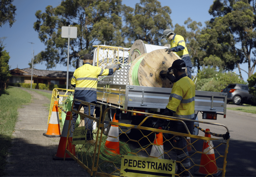 Three workers rolling out fibre cable from spool on the back of truck. A sign says 'pedestrians' with an arrow pointing left.