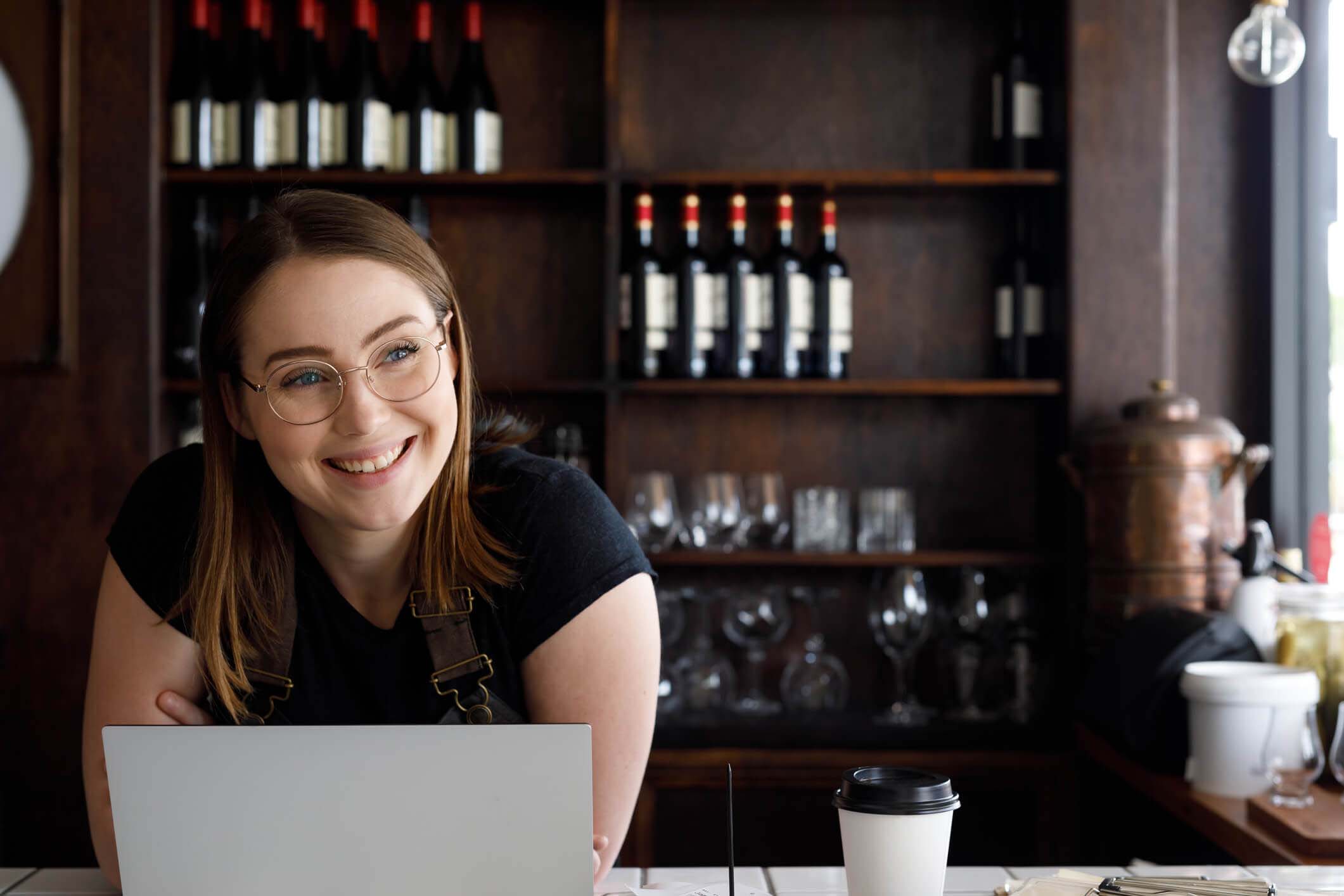 A young woman with a laptop in front of a selection of wines in a hospitality venue.