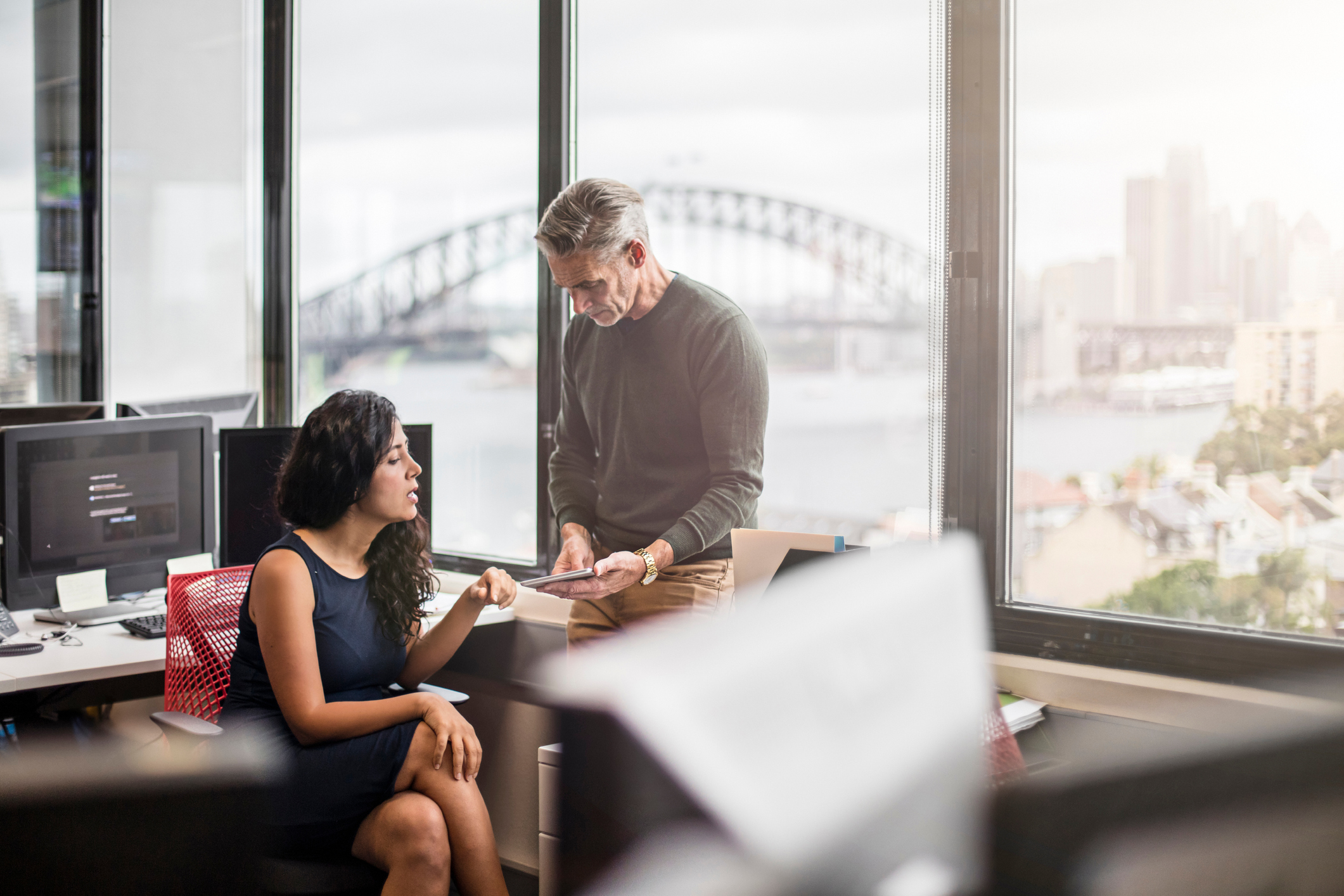 Image depicts an office setting with a man showing a woman information on a tablet.