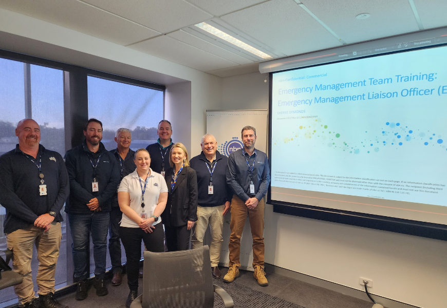 Eight smiling nbn Emergency Management Liaison Officers stand in front of a projector screen during a training session